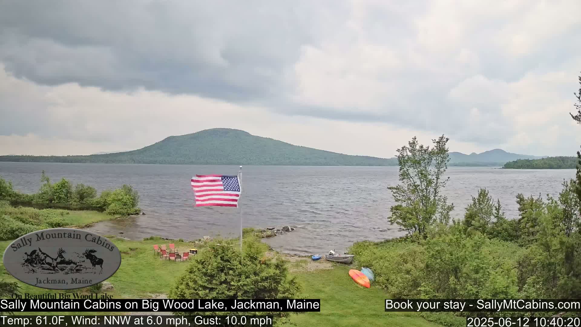 A calm lake is seen with a green shoreline, an American flag waving gently, and some kayaks and a small motorboat pulled ashore under an overcast sky.
