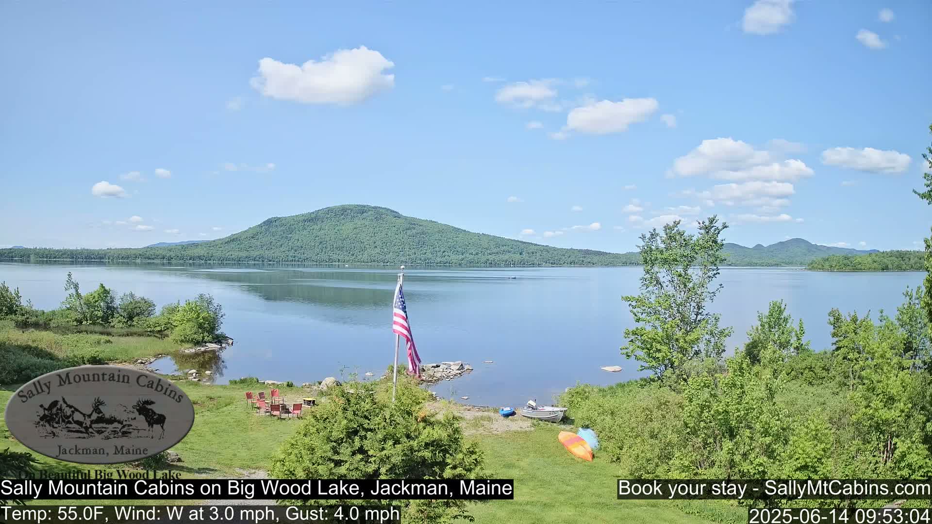 A calm lake with a forested mountain in the background, under a partly cloudy sky, is viewed from a lakeside property with a few chairs and a kayak.