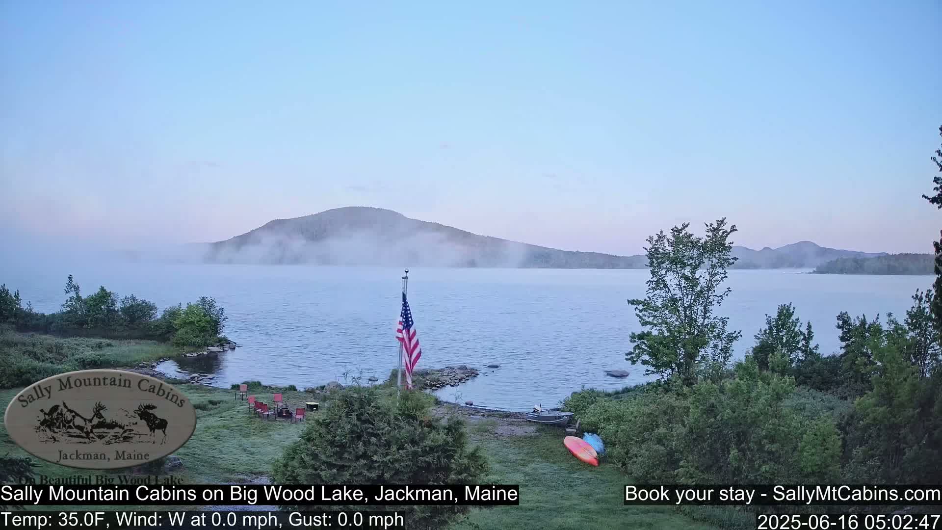 A calm lake with fog rolling in over the distant mountains, a small American flag planted near the shoreline, and kayaks resting on the grassy bank.
