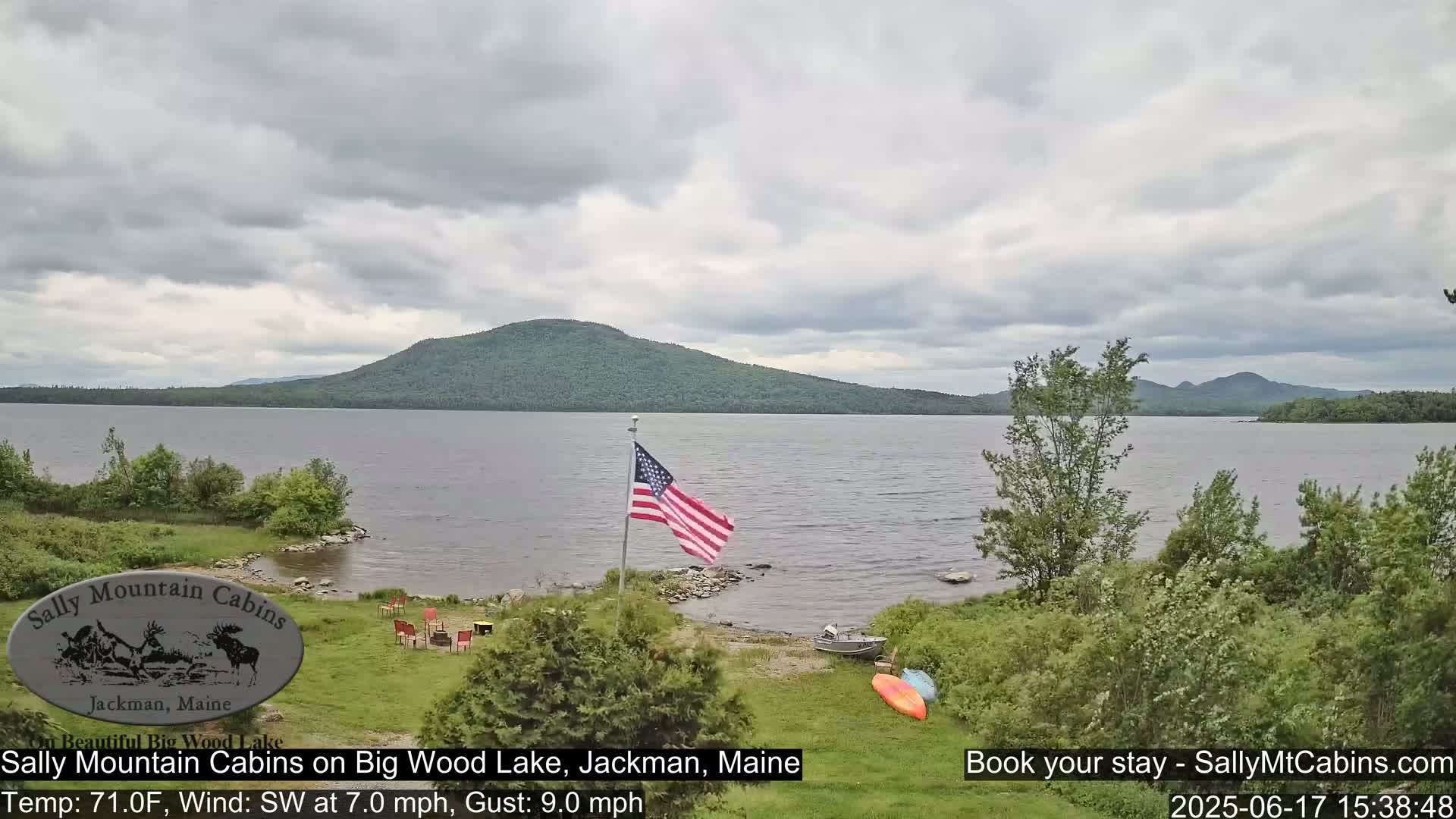 An American flag stands on the shore of a lake under a cloudy sky, with mountains in the distance and several kayaks and a small boat on the shoreline.
