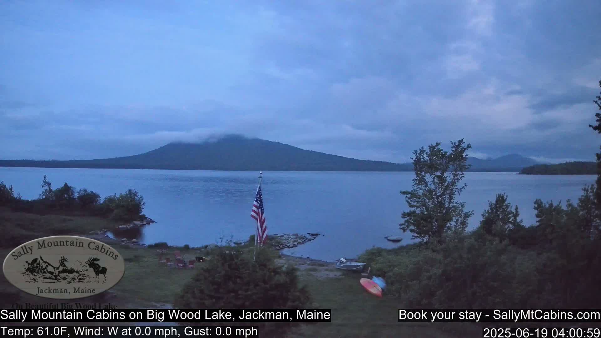 A calm, overcast evening scene shows a lake with a mountain in the distance, an American flag planted near the shoreline, and a small boat near the water's edge.