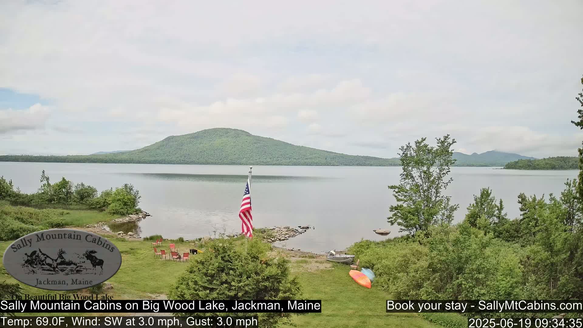 A calm lake with a wooded mountain in the background is viewed from a grassy lakeshore with a few red chairs, a small boat, and kayaks, under an overcast sky.