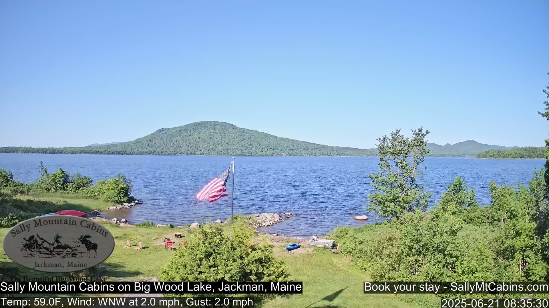 A calm, sunny day shows a view of a lake with a forested hillside in the background, a US flag, and some outdoor furniture on the grassy lakeshore.