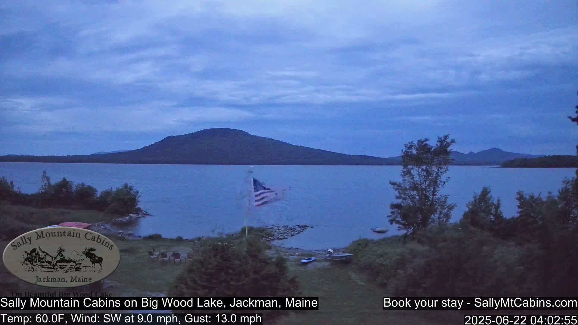 A calm lake at dusk, under a partly cloudy sky, is nestled between a shoreline with trees and a distant mountain range.