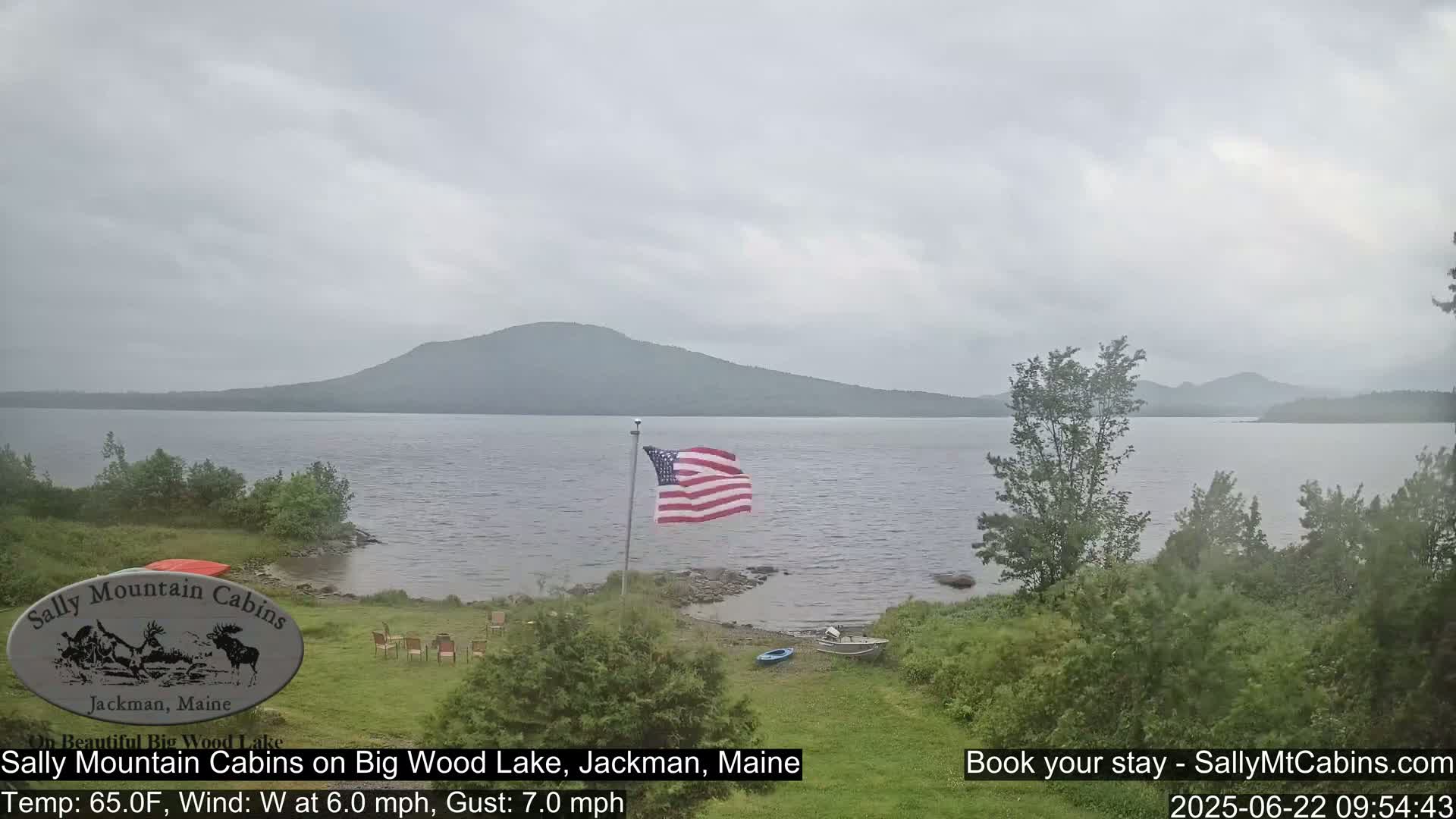 A calm lake with a distant mountain range under an overcast sky is viewed from a grassy lakeshore where an American flag is flying.