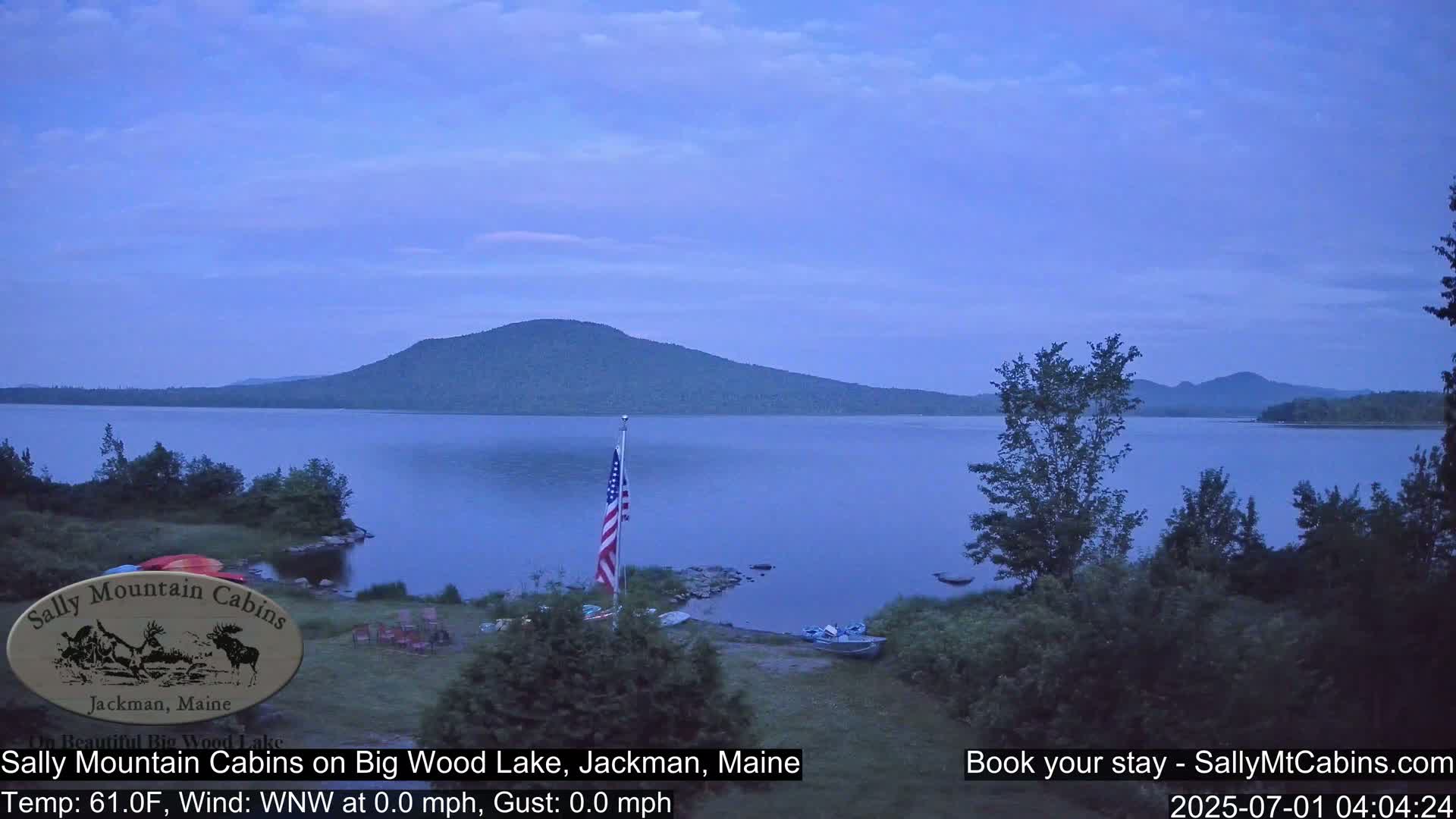 A calm, twilight view of a lake and distant mountains under a clear, dusky sky, with an American flag displayed near the shoreline.