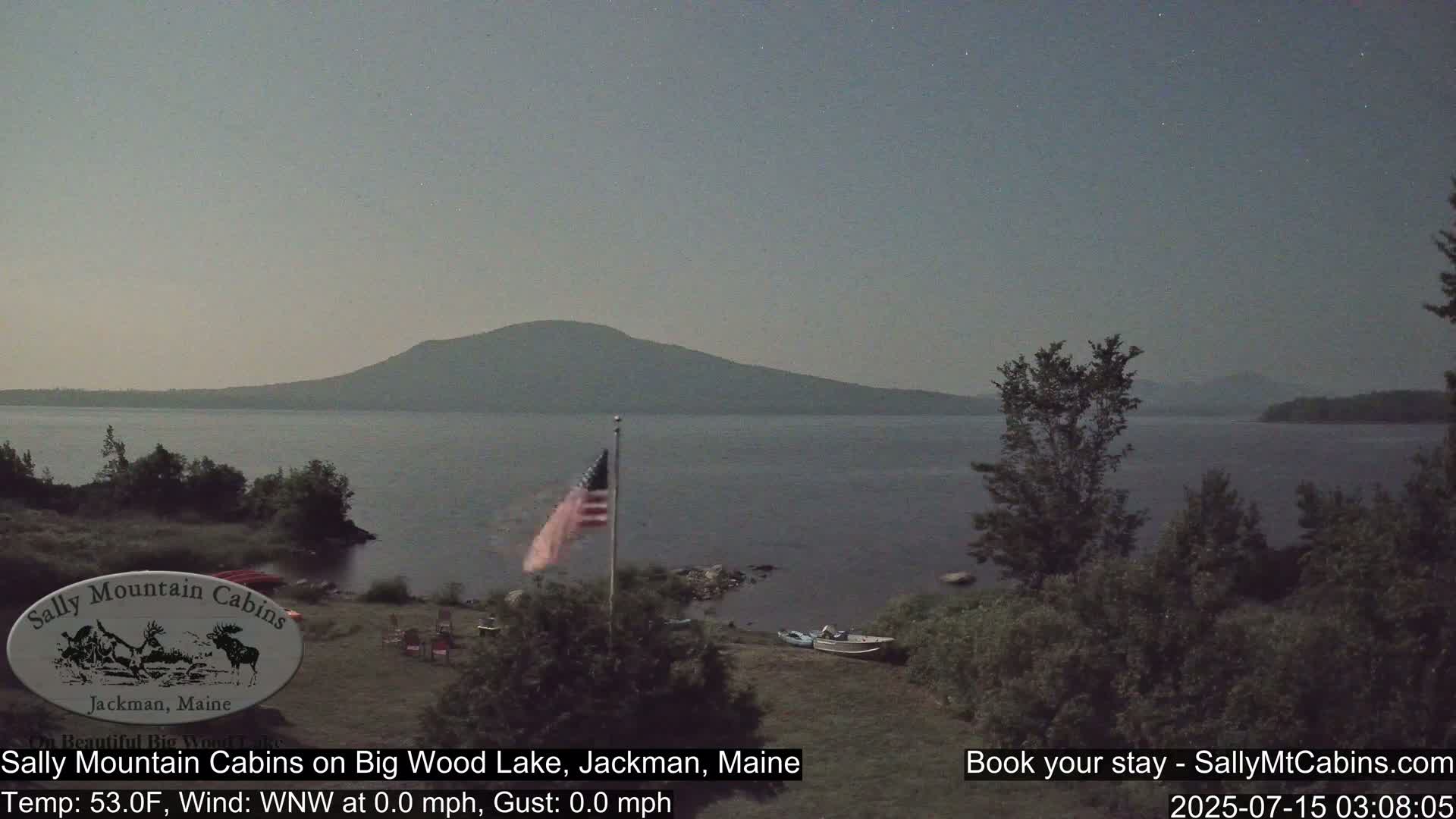 A calm, nighttime scene shows a lake with a distant mountain range under a dusky sky, a flagpole with an American flag, and a small boat near the shoreline.