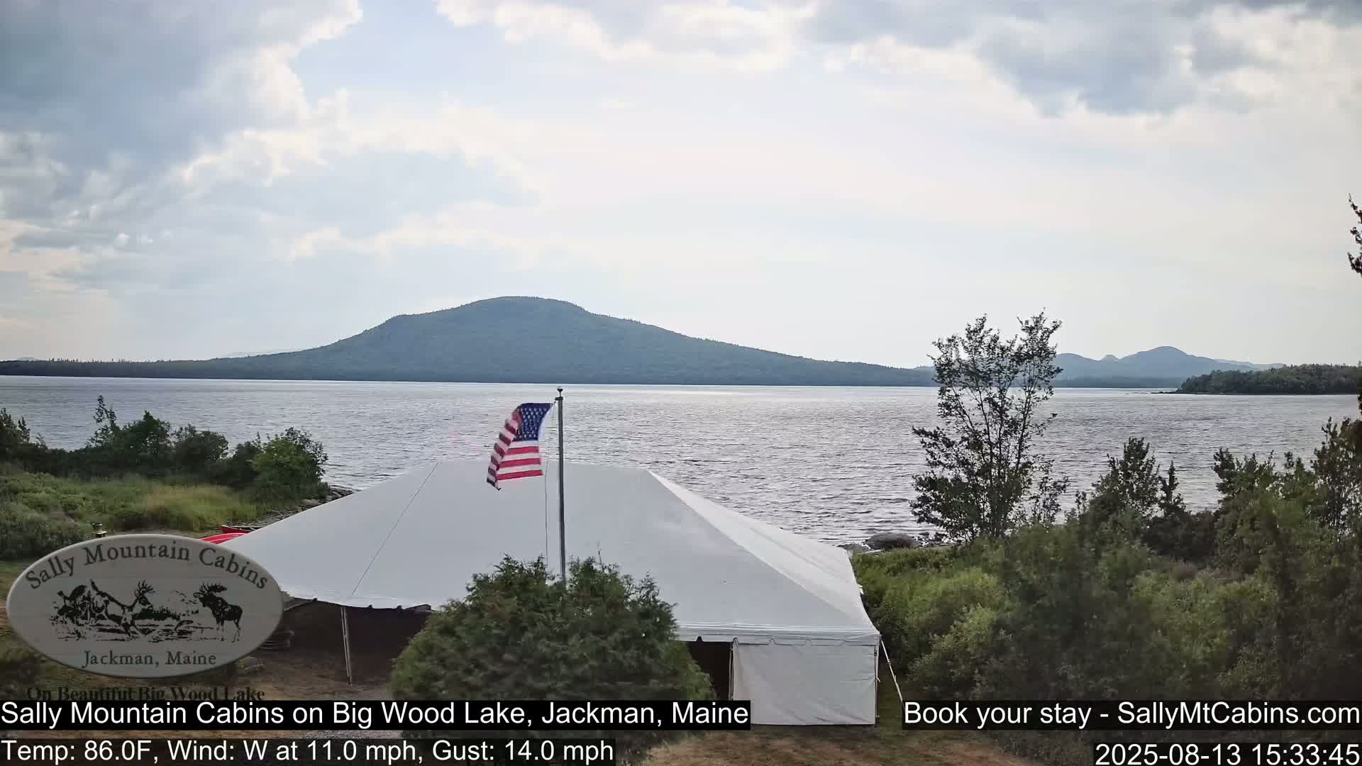 A large white tent sits near a calm lake with a mountain range in the background under a partly cloudy sky with a gentle west wind.
