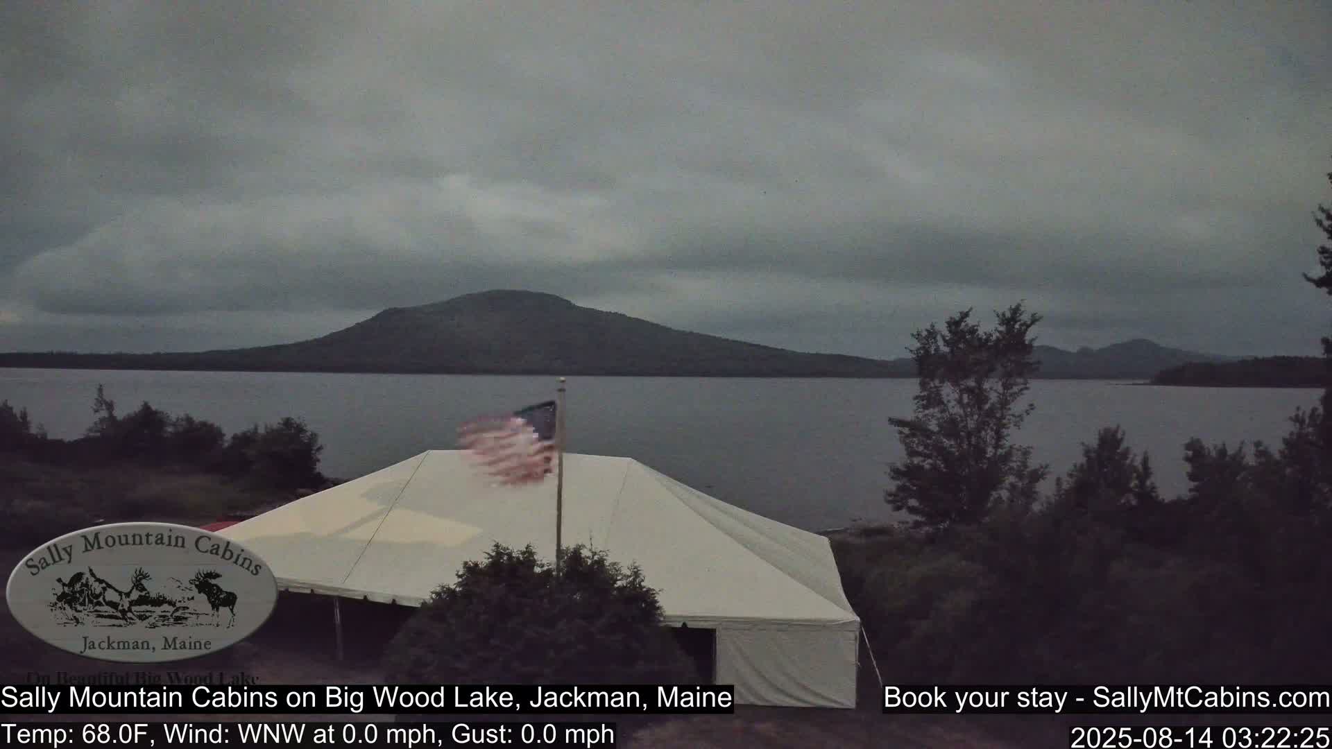 A large white tent sits near a calm lake under an overcast sky, with a distant mountain visible and an American flag waving gently in the light breeze.