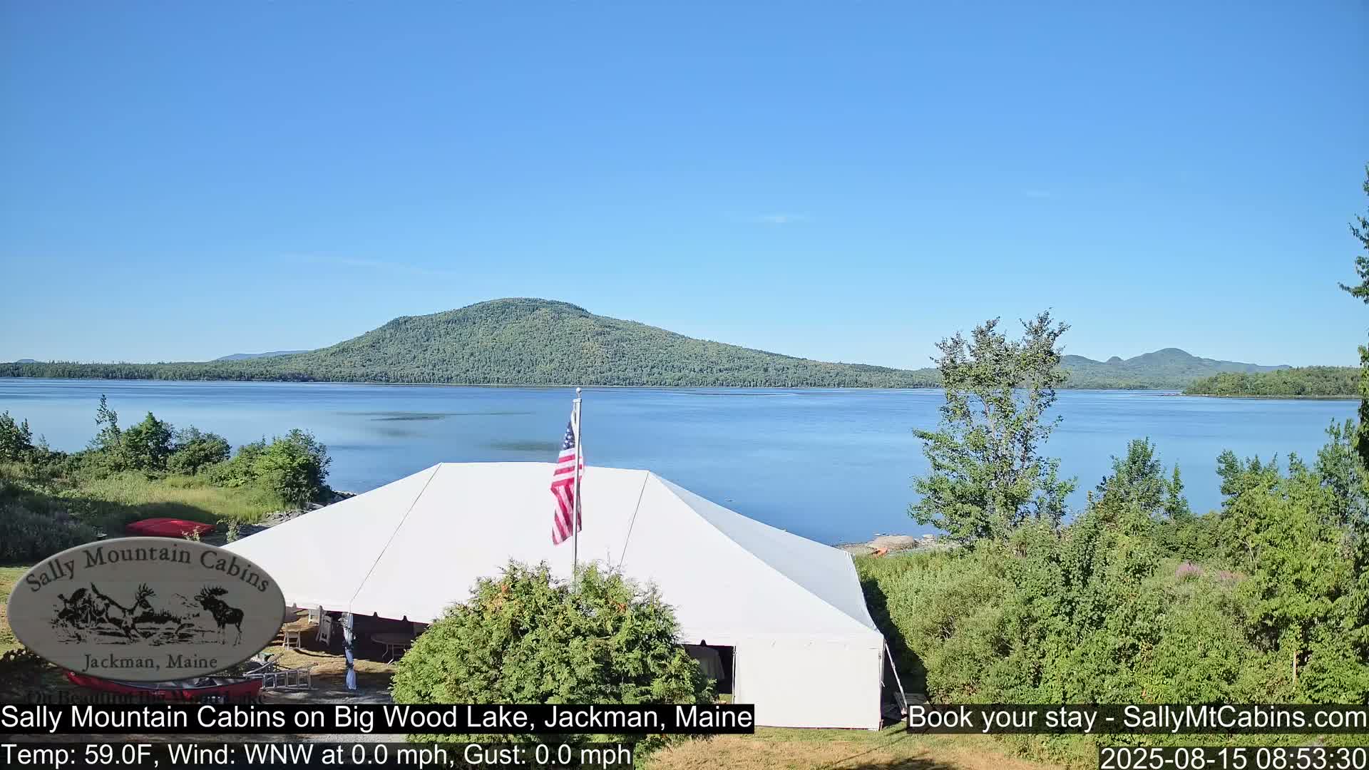 A large white tent sits near a calm lake under an overcast sky, with a distant mountain visible and an American flag waving gently in the light breeze.