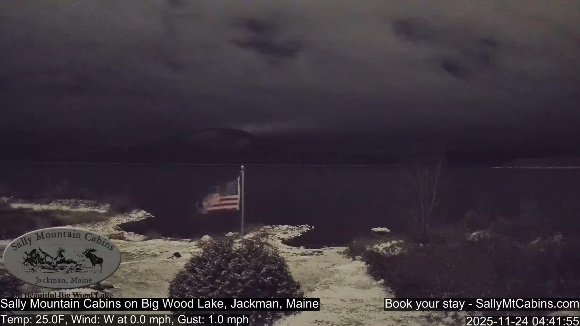 An American flag stands on a snow-dusted shore next to a dark lake under an overcast sky, with silhouettes of distant mountains visible through the dim, cold morning light.
