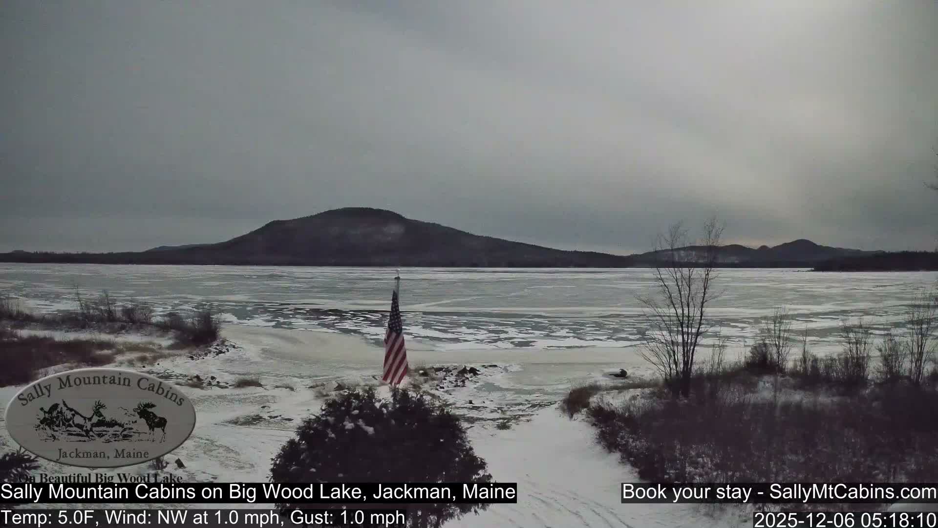 A wide, wintry scene shows an American flag planted on a snow-covered bank beside a partially frozen lake, with dark, tree-covered mountains lining the horizon under a dull, overcast sky.