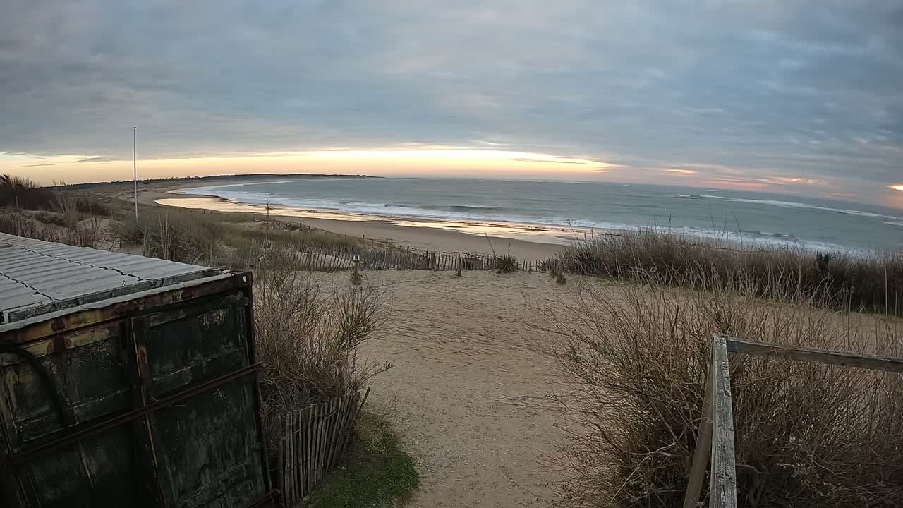 A sandy beach with gentle waves under a partly cloudy sunset sky is viewed from behind a weathered container and wooden fence.