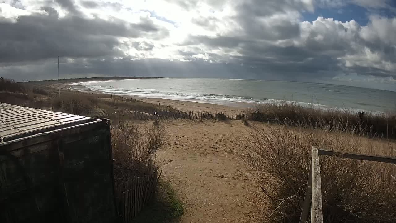 A sandy beach with low, brown vegetation slopes down to a somewhat choppy ocean under a cloudy sky.