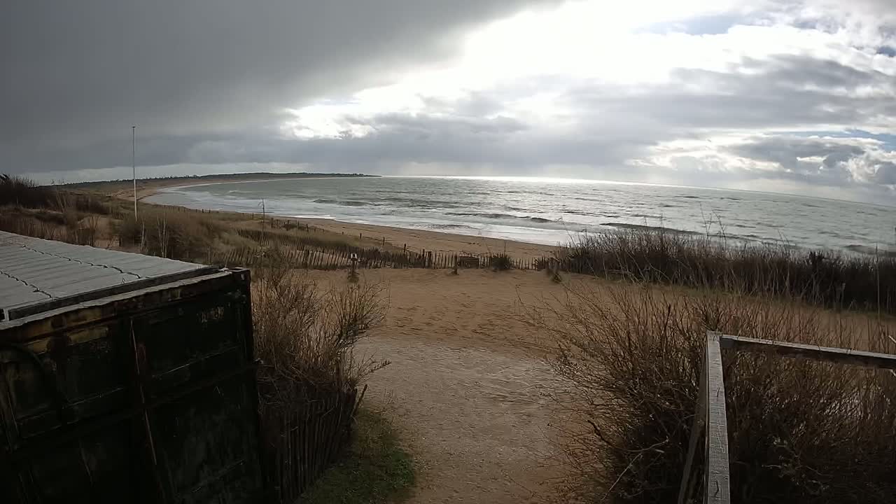 A sandy beach with a moderately wavy ocean under a partly cloudy, gray sky is seen from behind a dune with dry, brown vegetation.