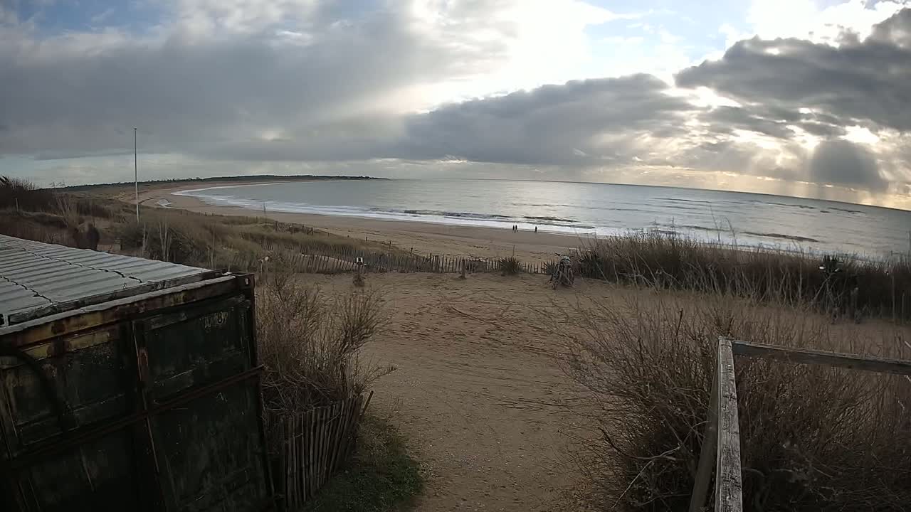 A cloudy but bright day at a beach with a sandy shoreline, gentle waves, and a metal container partially visible in the foreground.