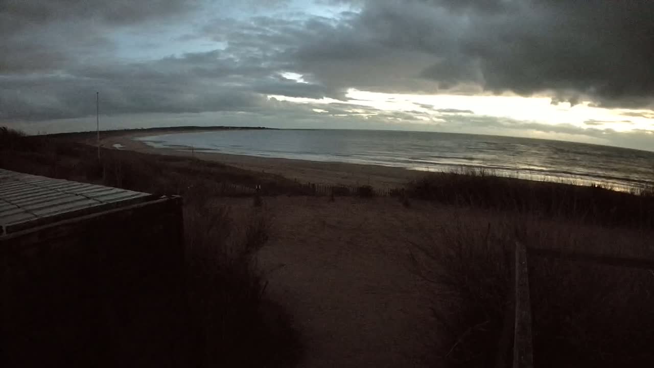 A view from a dune overlooking a beach and a calm sea under a mostly cloudy, dark sky with hints of sunset.
