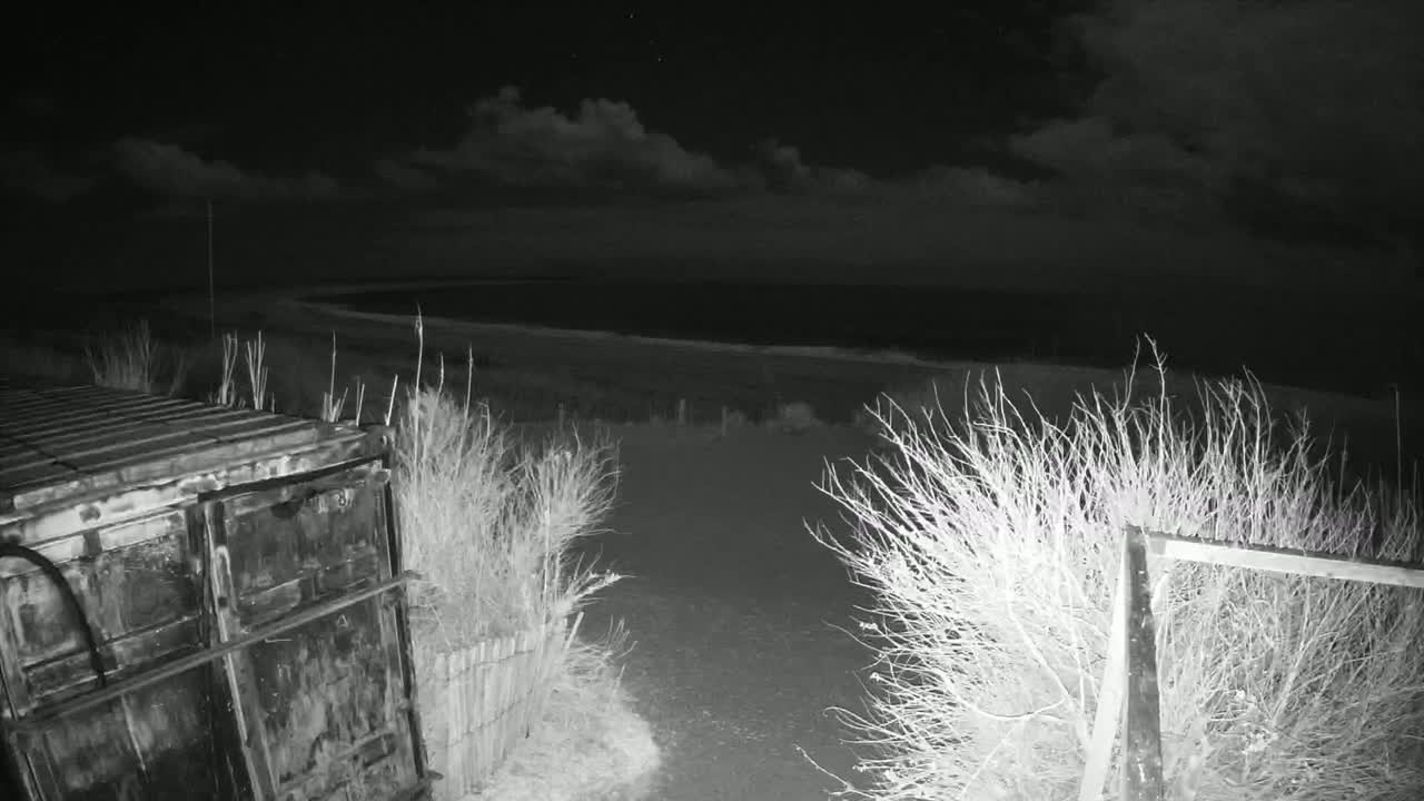 A nighttime, low-light image shows a dark, metal container next to a dirt path leading to a body of water under a cloudy sky.