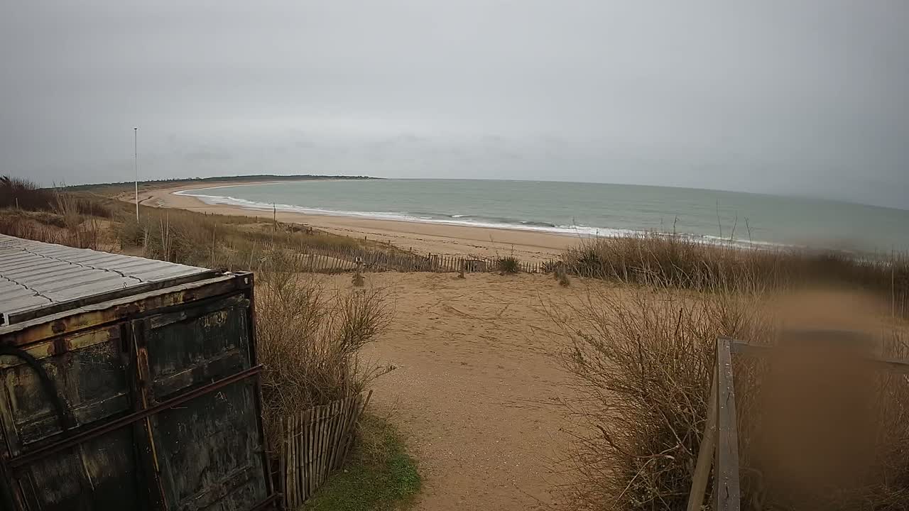 A weathered container sits near a sandy path overlooking a beach with gentle waves under an overcast sky.