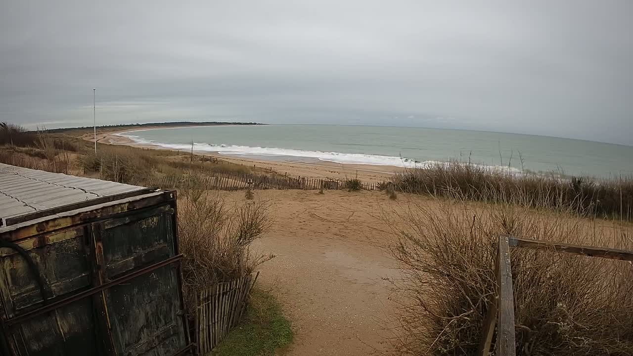 A sandy beach with gentle waves under an overcast sky is viewed from behind a weathered container and low fence.