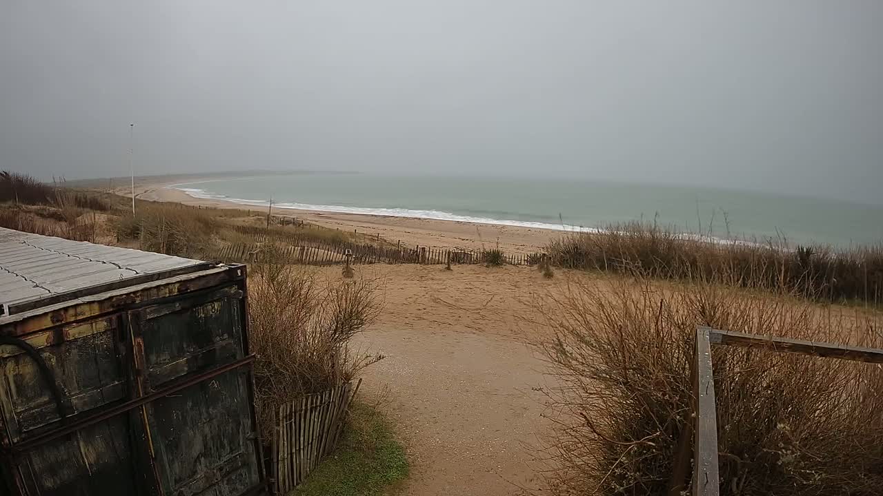 A weathered metal container sits near a sandy path leading to a beach with gray, overcast skies and gentle waves.