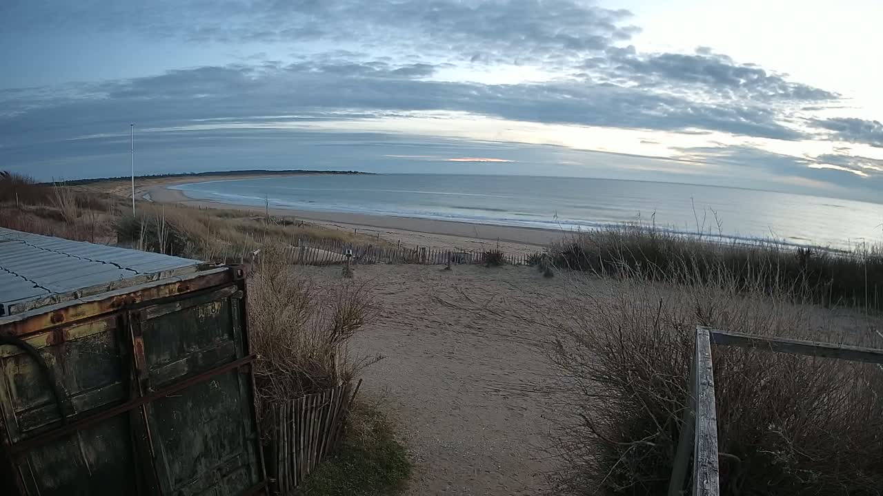 A rusty container sits atop a dune overlooking a calm, gray ocean under a partly cloudy sky.