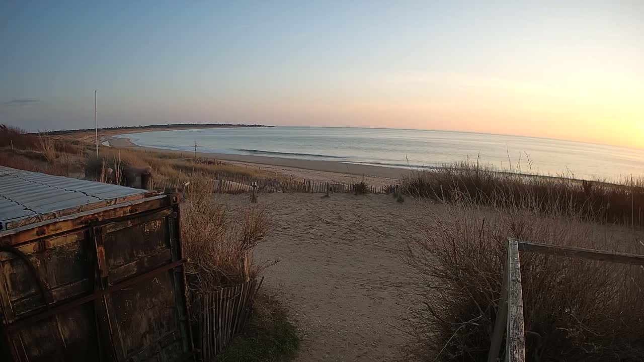 A rusty container sits near a sandy path overlooking a calm ocean beach at sunset.