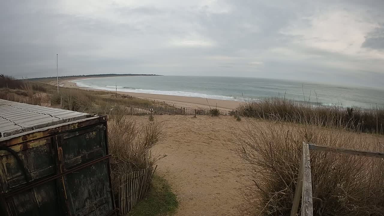 A weathered container sits atop a sandy dune overlooking a grey, overcast ocean with small waves lapping a wide sandy beach.