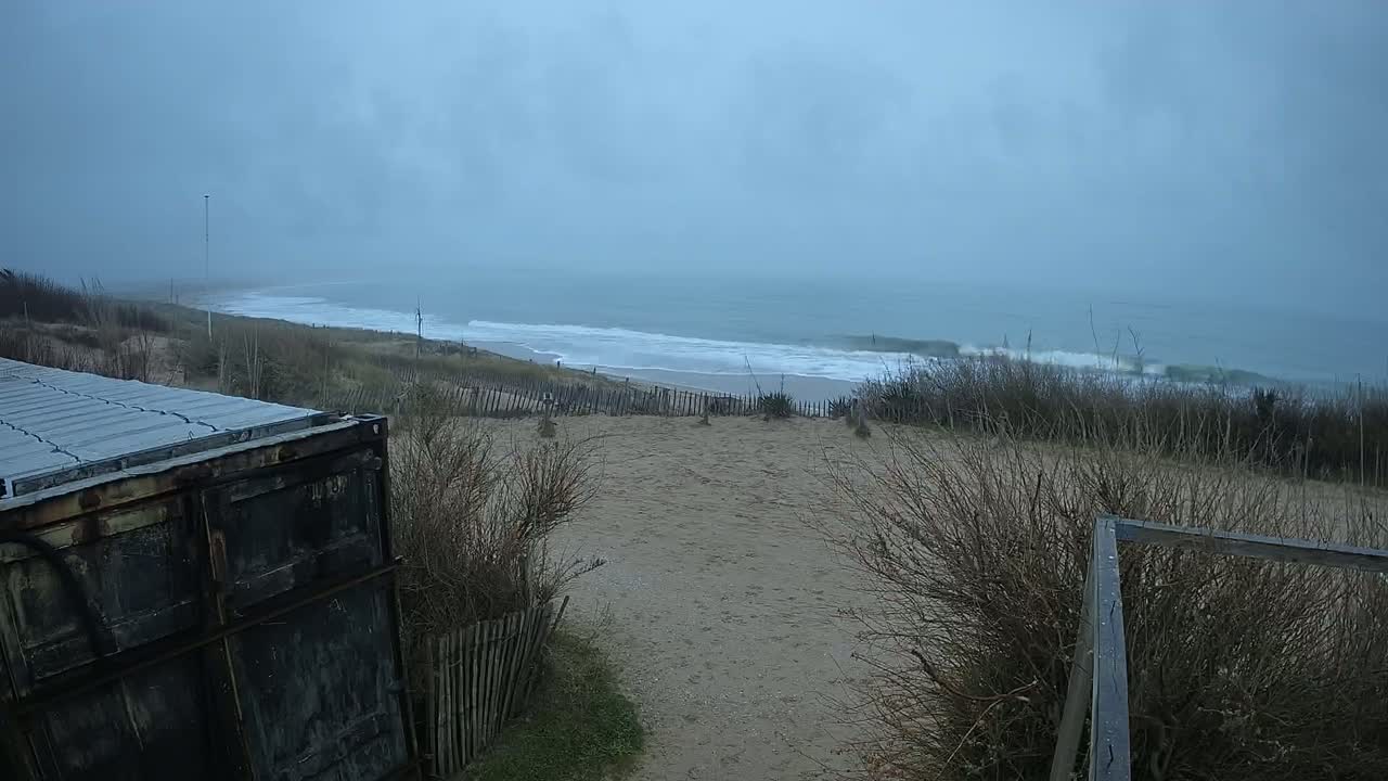 A dark gray, overcast sky hangs over a beach with moderate waves, viewed from behind a weathered container and dune vegetation.
