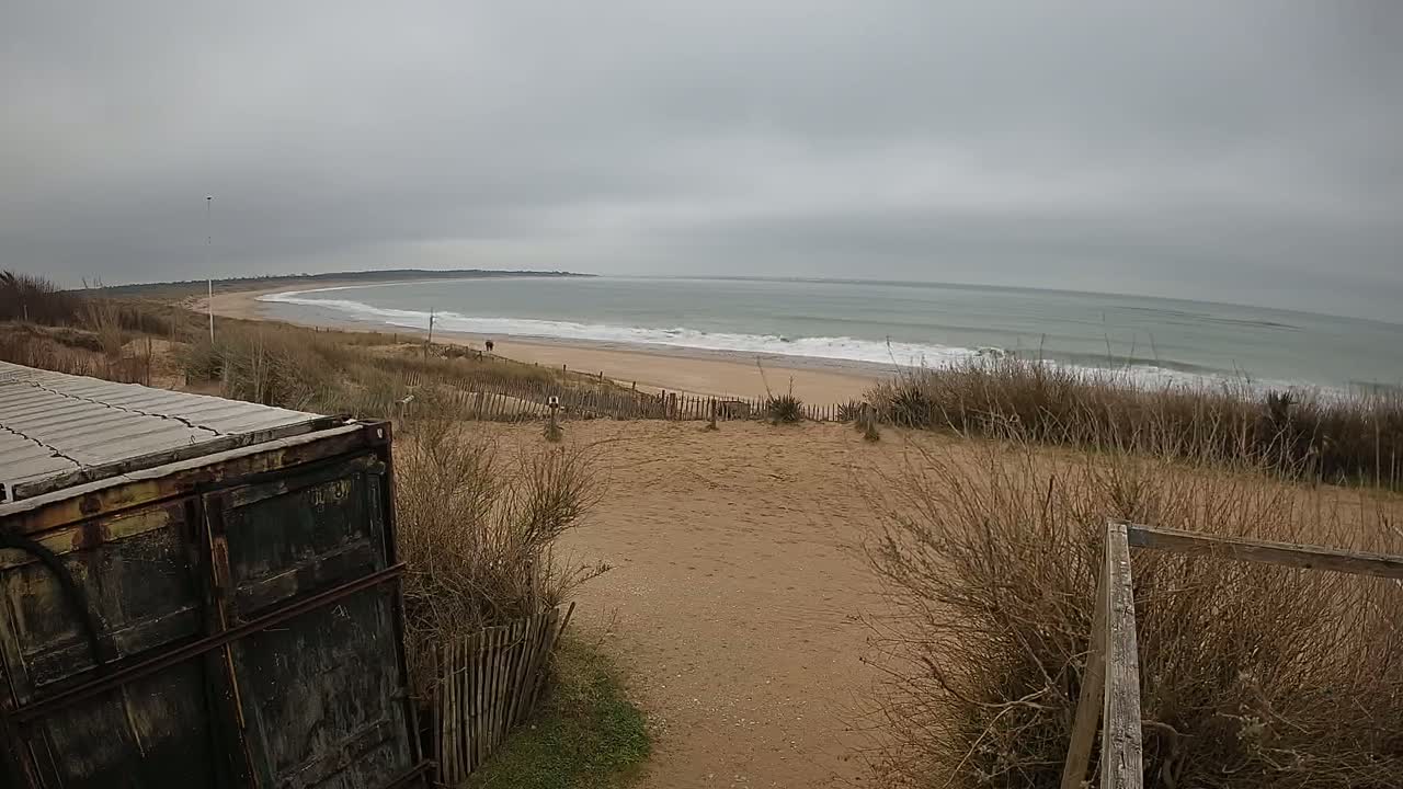 A weathered container sits partially visible in the foreground, overlooking a sandy beach with gentle waves under a cloudy sky.