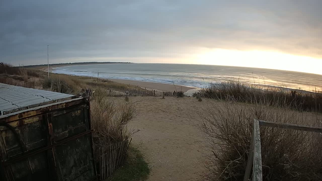 A sandy path leads down to a beach with waves under a cloudy but brightening sky.