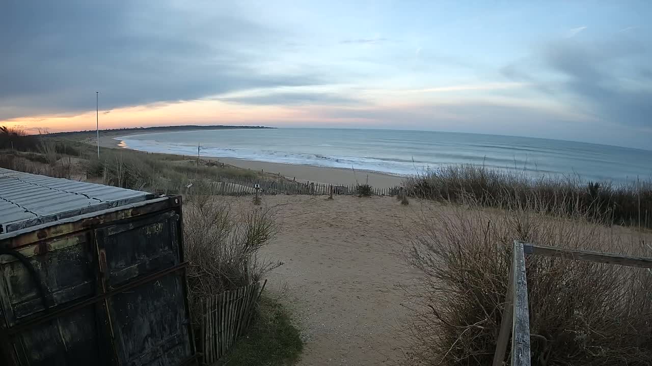 A weathered shipping container sits beside a sandy path overlooking a beach with gentle waves under a partly cloudy sunset sky.