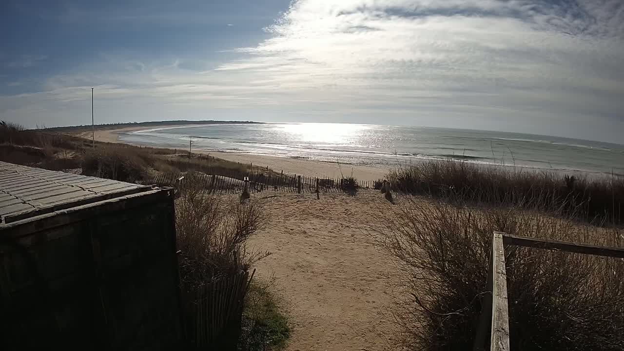 A sandy beach path leads to a calm ocean under a partly cloudy sky.