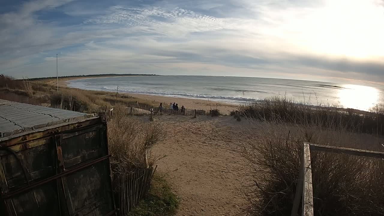 A sandy beach with gentle waves under a partly cloudy sky, a group of people visible in the distance, and a weathered container in the foreground.