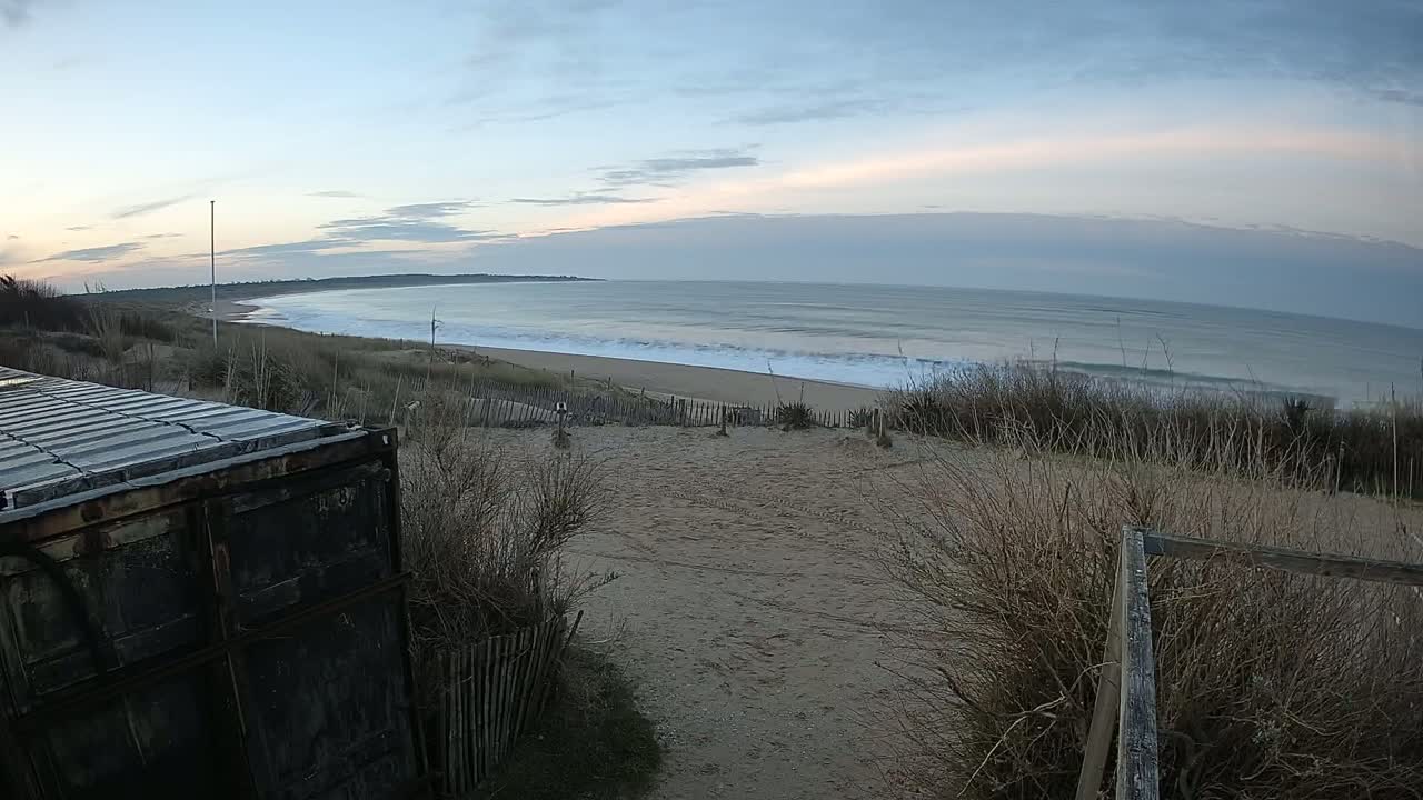 A dark-colored container sits near a sandy path overlooking a beach with gentle waves under a mostly cloudy, pale sky.