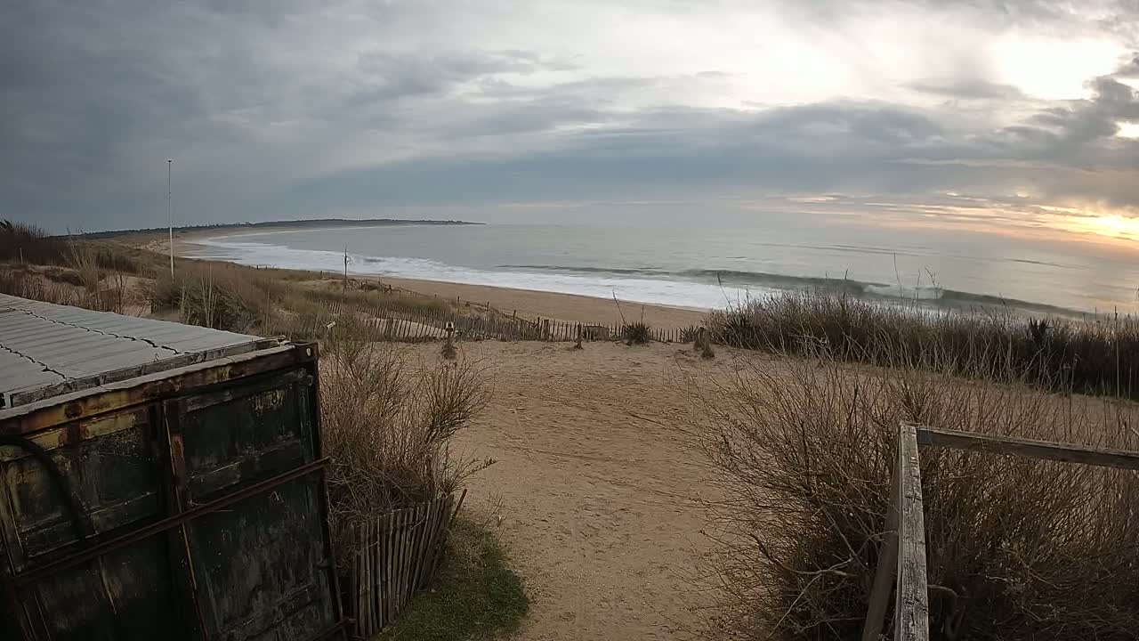 A weathered container sits near a sandy dune overlooking a beach with gentle waves under a cloudy sky.