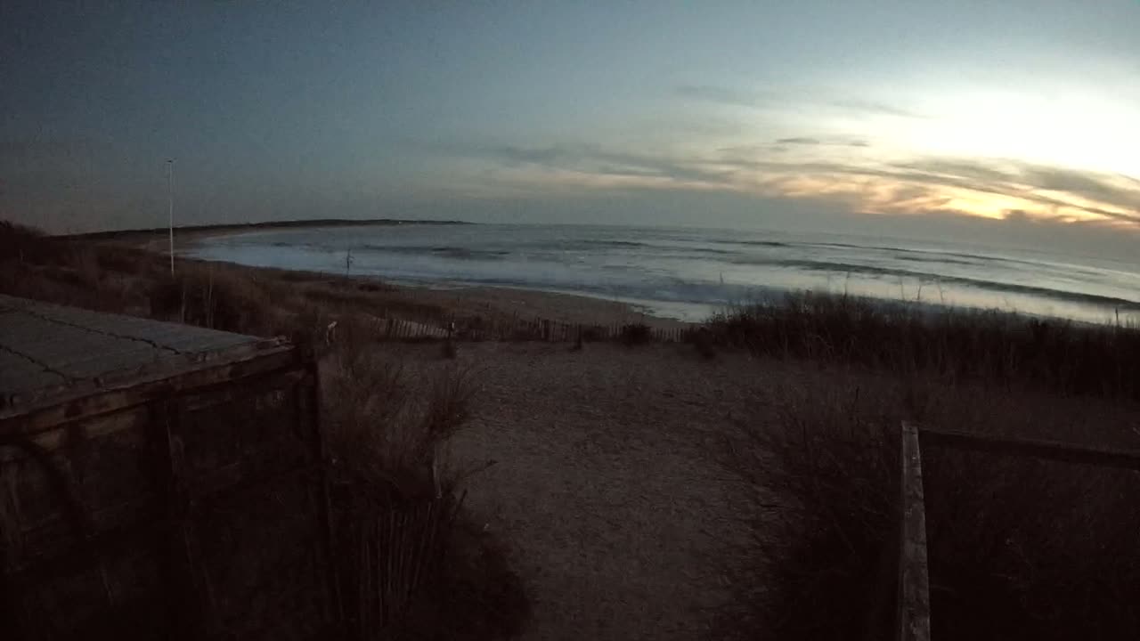 A view from a dune overlooking a beach with gentle waves under a dusky sunset sky.