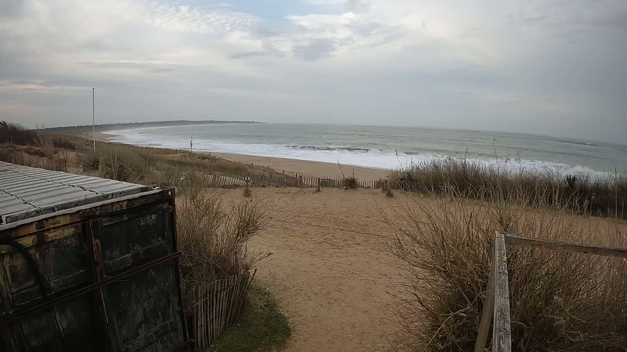 A view from behind a weathered container shows a sandy beach with small waves under an overcast sky.