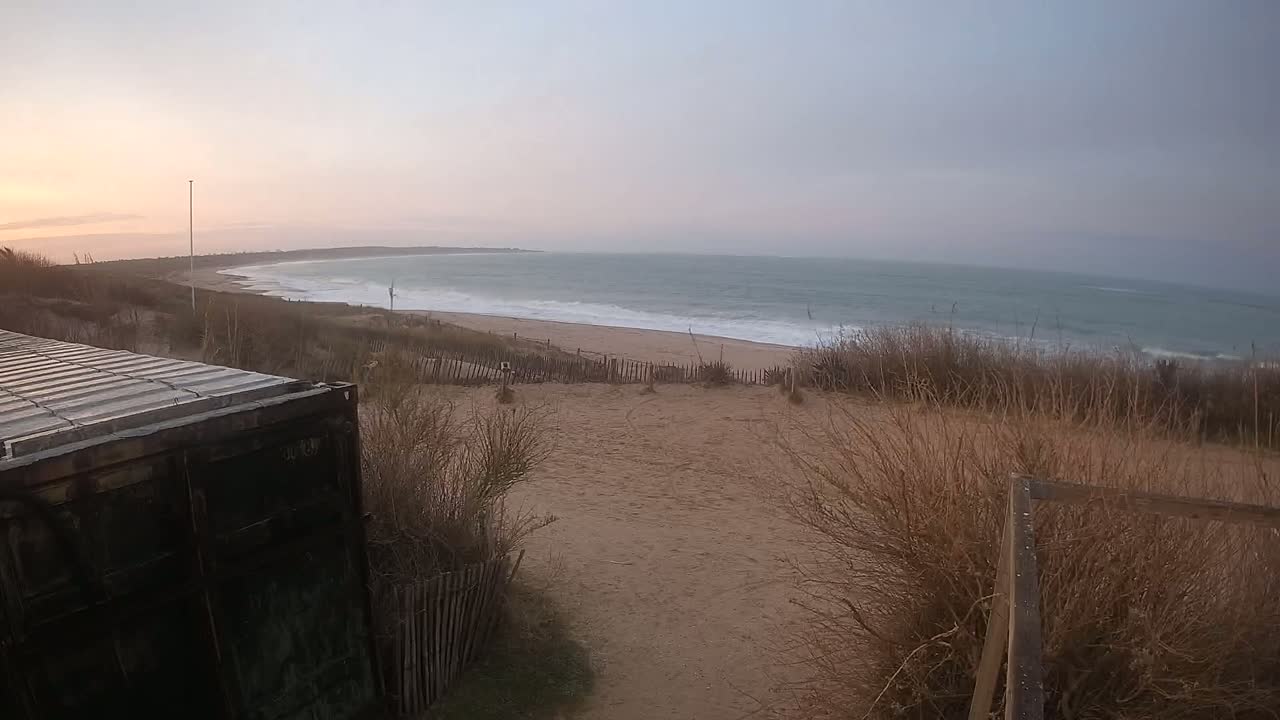 A dark shipping container sits beside a sandy dune overlooking a beach with gentle waves under a cloudy sunset sky.