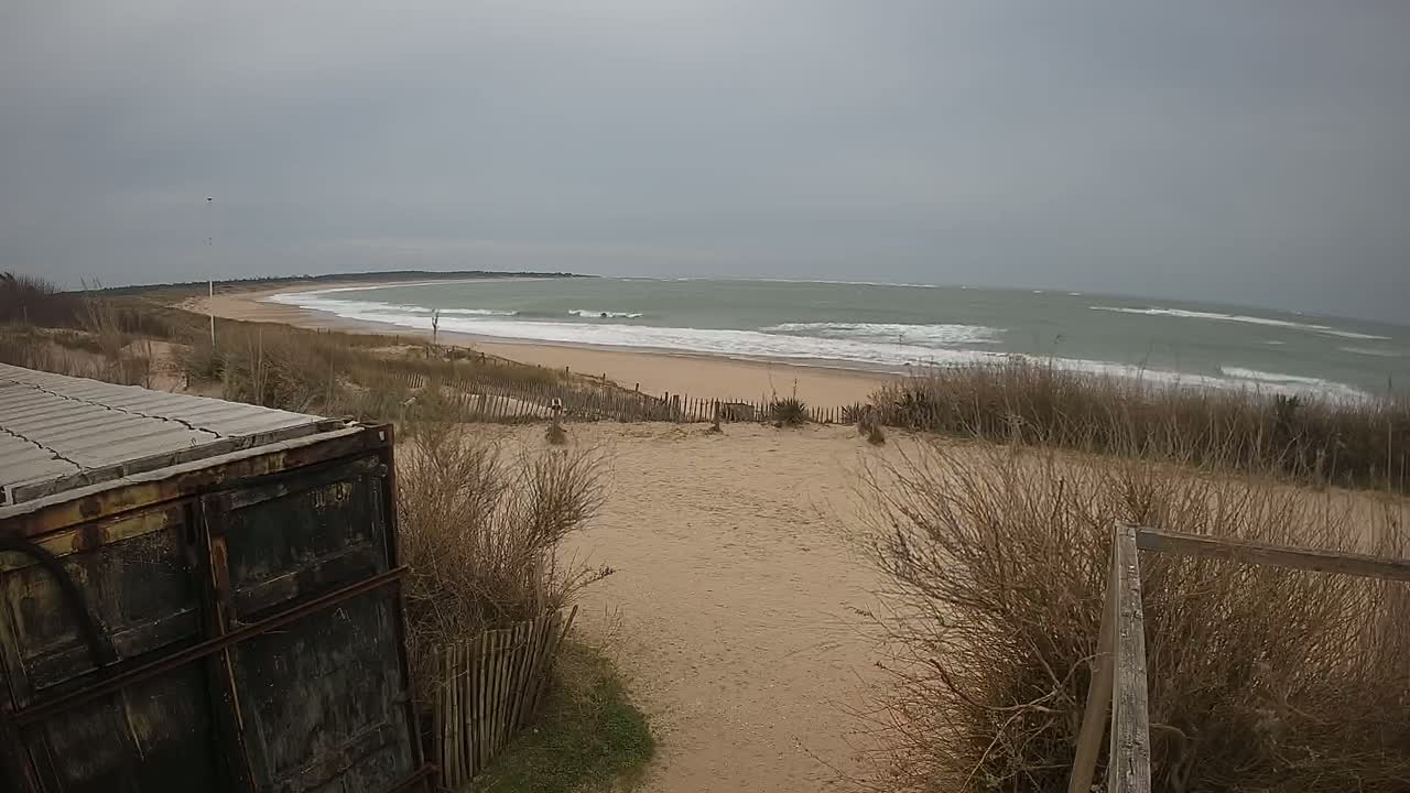A sandy beach with moderate waves under an overcast sky is seen from behind a weathered container of building materials and a dune-top fence.