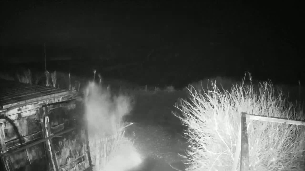 A nighttime, low-light image shows a dark, possibly metallic structure emitting a cloud of dust or smoke near sparse brush and a metal fence.