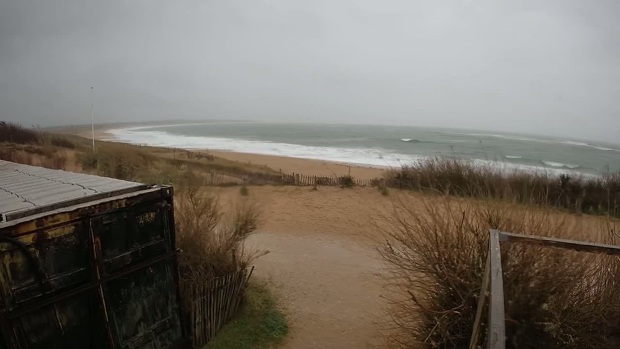 A stormy gray day reveals a view of a beach with waves, sand dunes, and scrub vegetation, partially obscured by a weathered metal container and a wooden fence.