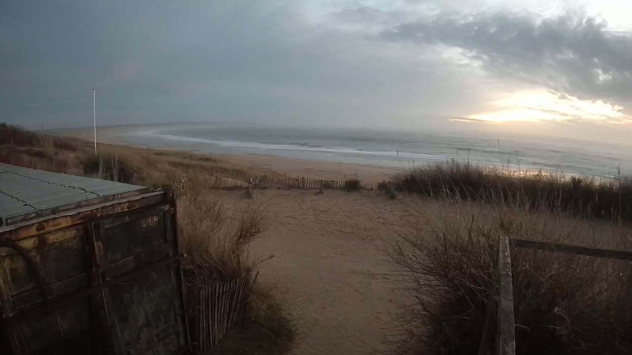 A sandy beach with gentle waves under a partly cloudy sky, viewed from behind a weathered wooden structure and dune grasses.
