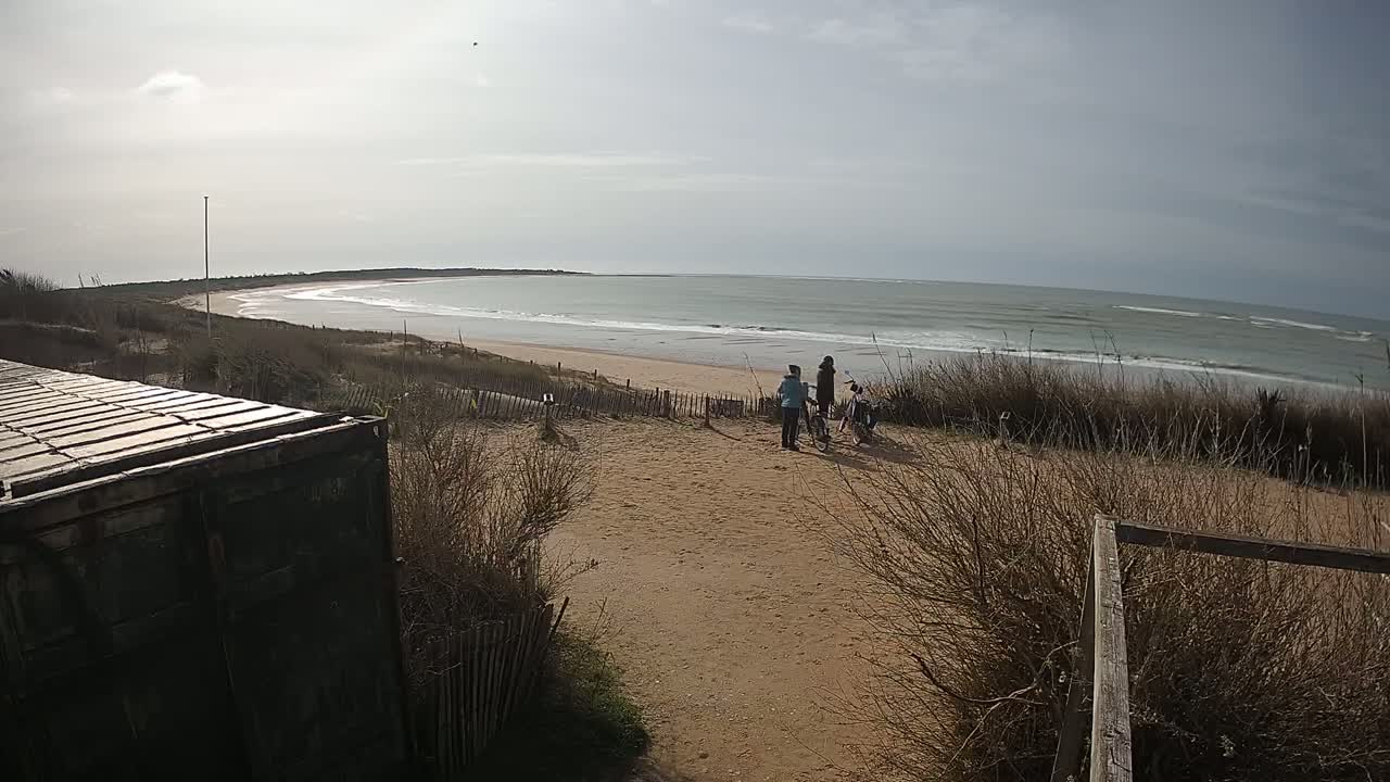 Three people stand near bicycles on a sandy dune overlooking a beach with gentle waves under a partly cloudy sky.
