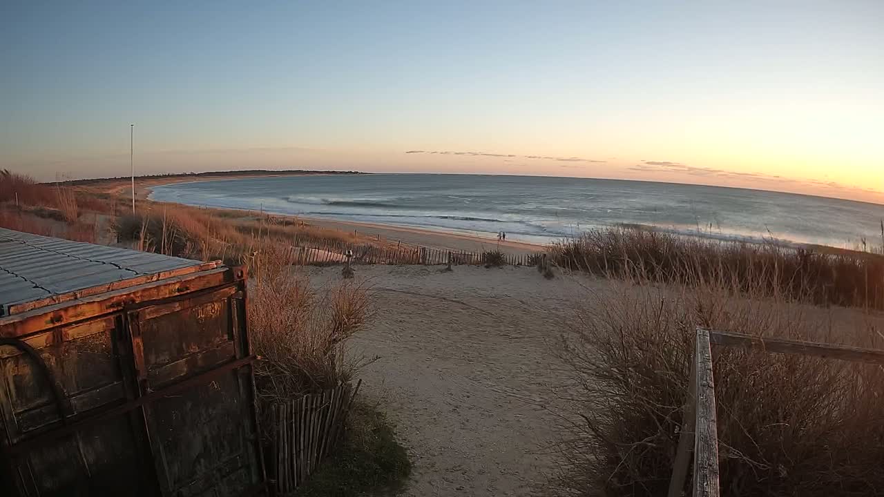 A sandy beach with gentle waves under a sunset sky is viewed from behind a weathered container and a dune with sparse, brown vegetation.