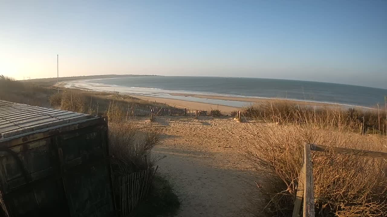 A sandy path leads down to a calm ocean beach under a clear, sunny sky, partially obstructed by dry brush and a dark metal container.