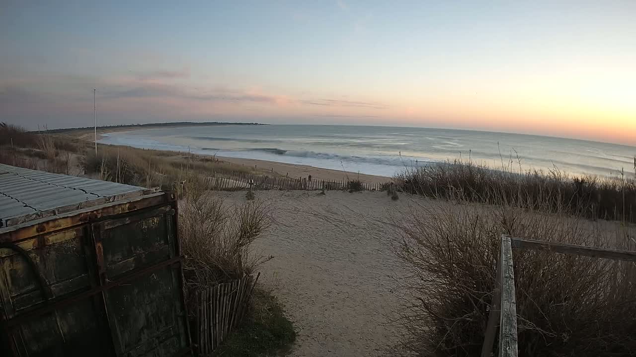 A sandy beach with gentle waves under a calm sunset sky is viewed from behind a weathered metal container and wooden fence.