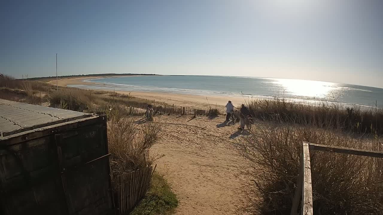 A sandy beach with calm ocean waves under a bright, sunny sky is viewed from behind a weathered metal container, with several people walking on the beach in the distance.