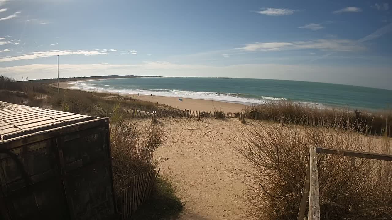 A sandy beach with a calm ocean under a mostly sunny sky is viewed from behind a weathered metal container and a wooden fence.