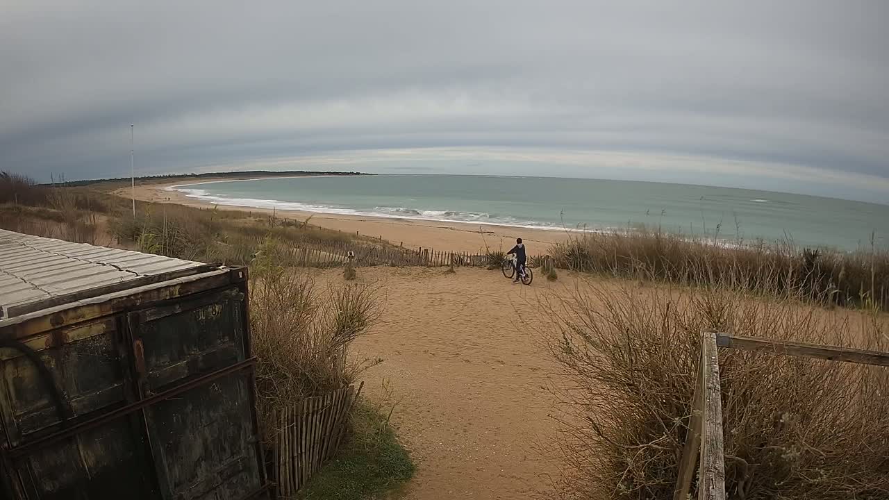 A person on a bicycle stands on a sandy beach near a weathered metal container, overlooking a calm, gray ocean under an overcast sky.